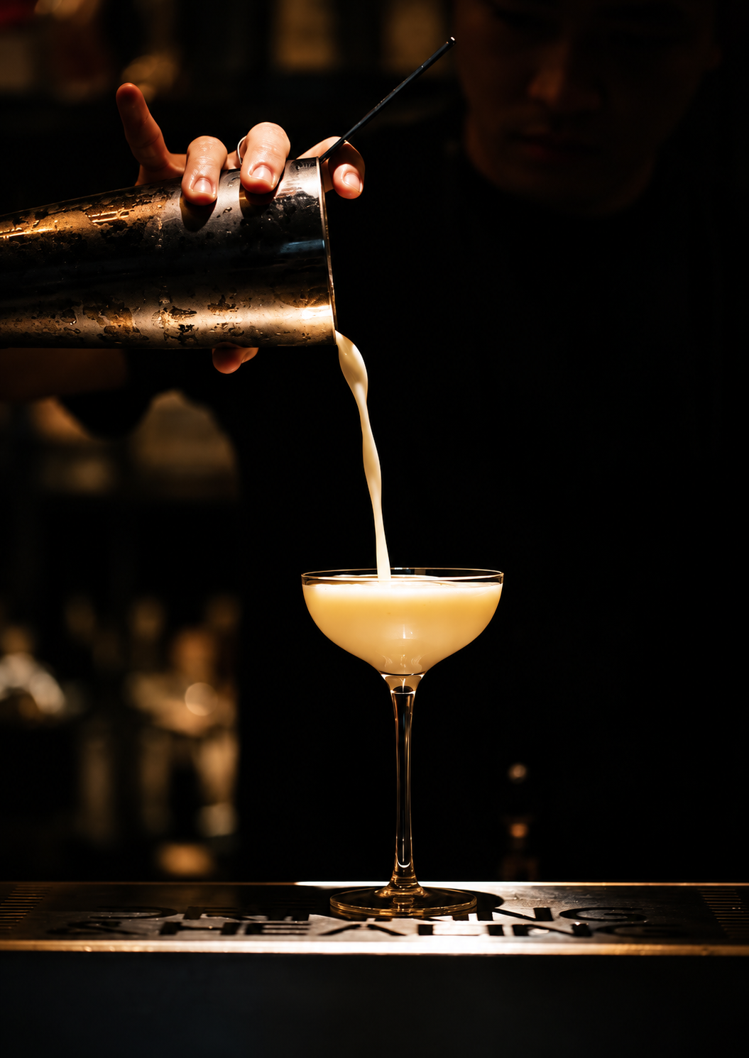 A creamy drink poured from a metal shaker into a coupe glass against a dark bar background.