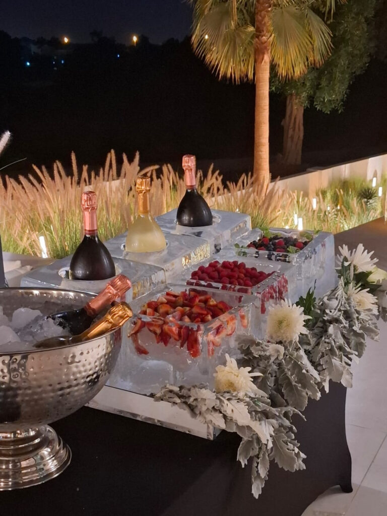 Champagne bottles and berries displayed in a sculpted ice station with white flowers at an outdoor evening event.