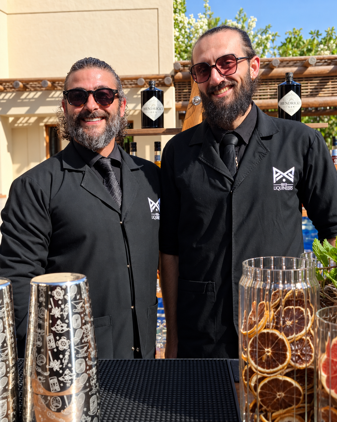 Tareq and Yamam smiling behind an outdoor bar in LiquiNeers uniforms, with cocktail tools and dried citrus garnishes