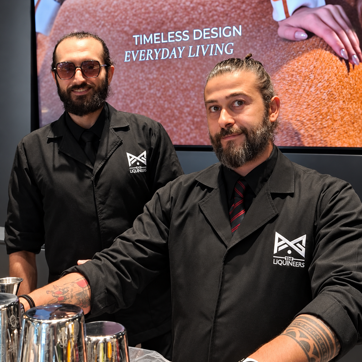 Tareq and Yamam in black LiquiNeers uniforms behind a bar with cocktail shakers.
