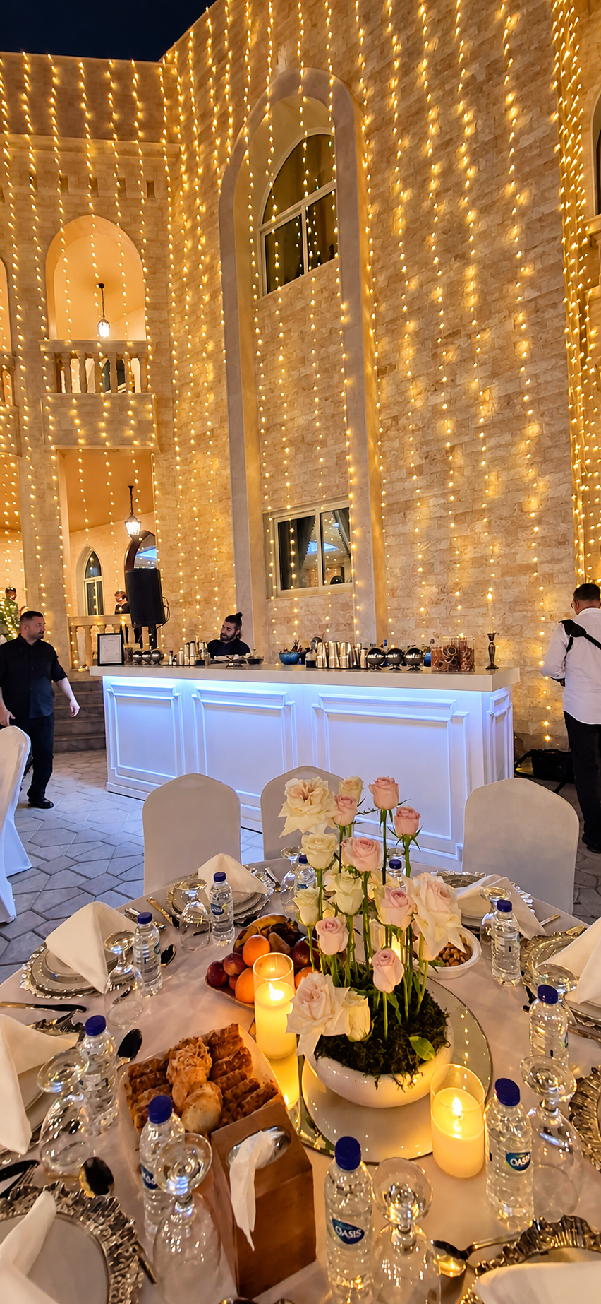 Illuminated white bar with mixologists beneath fairy lights at an evening celebration