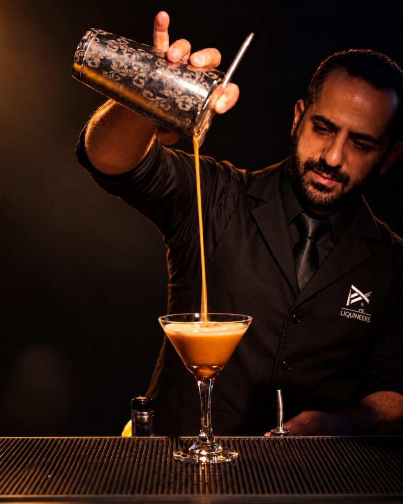 Ali pouring an amber cocktail while wearing a black LiquiNeers vest and tie