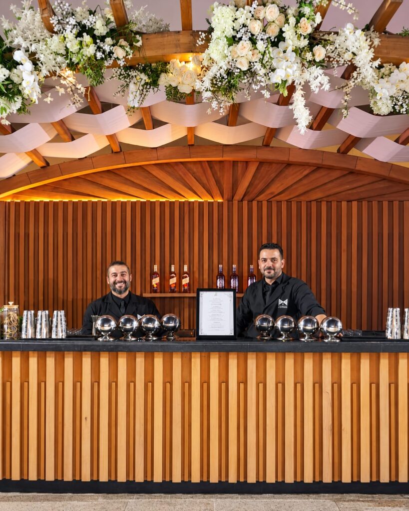 Two LiquiNeers mixologists behind a wooden bar beneath white flowers