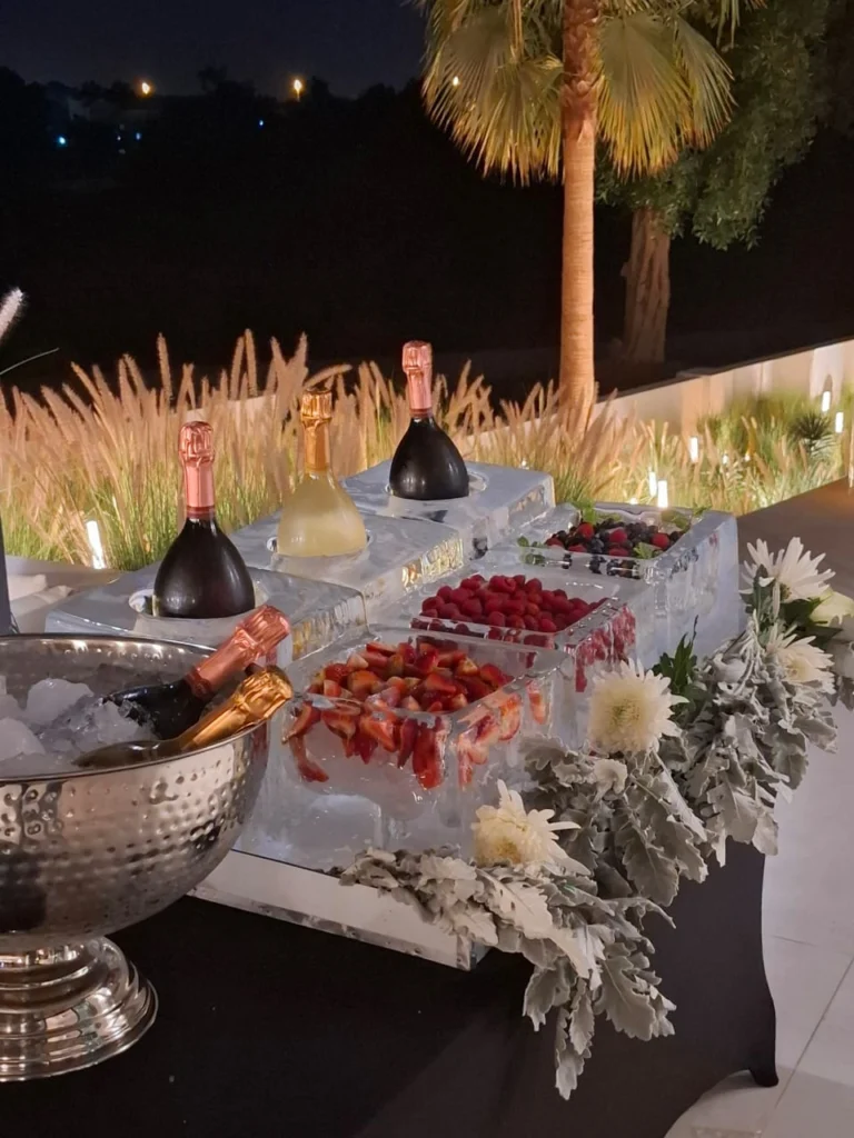 Champagne bottles and berries displayed in a sculpted ice station with white flowers at an outdoor evening event.