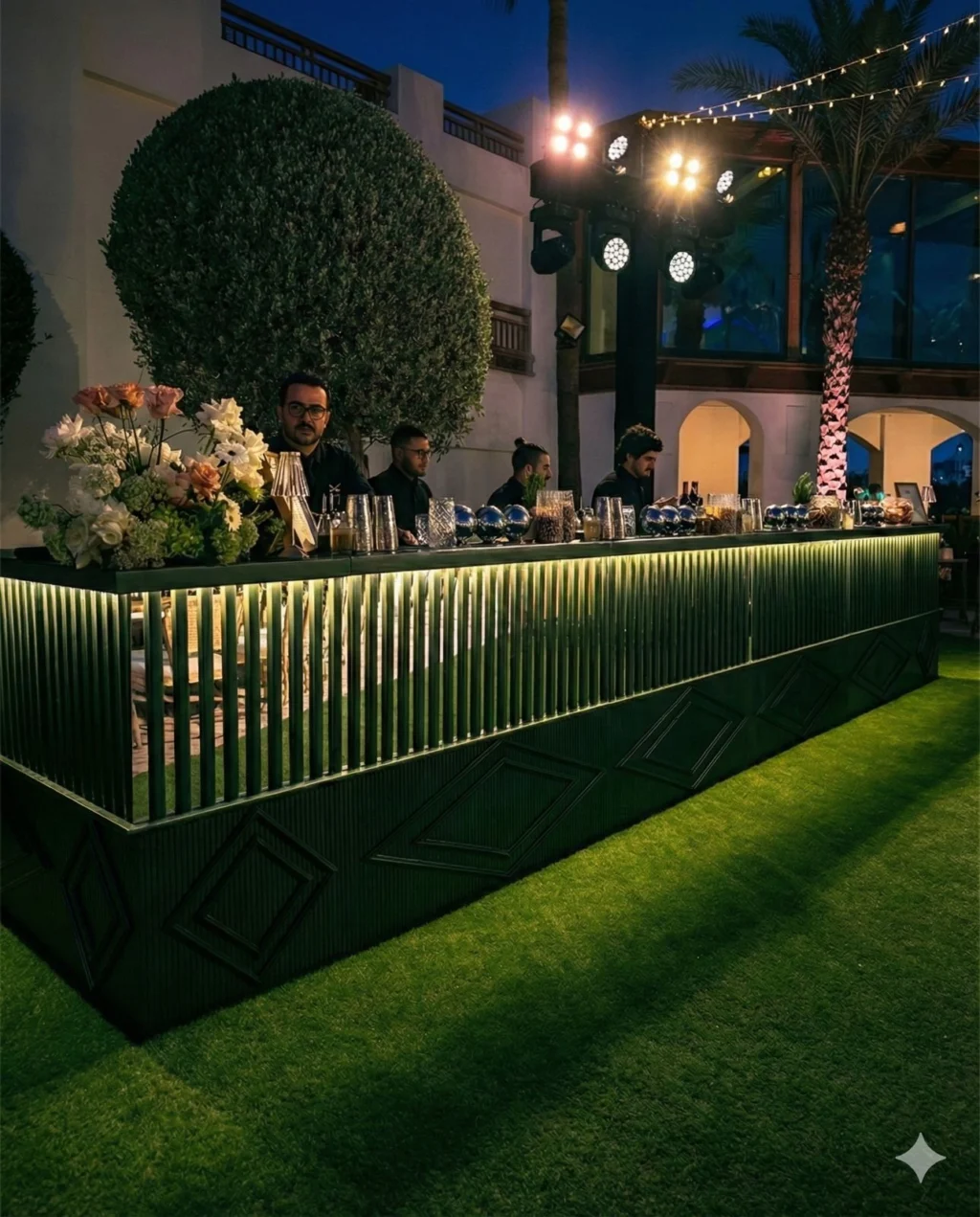 LiquiNeers mixologists behind an illuminated outdoor bar with glassware, cocktail tools and flowers.