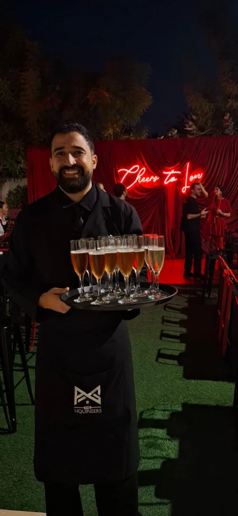 Waiter in a black LiquiNeers apron carrying a tray of flute glasses at an outdoor evening event