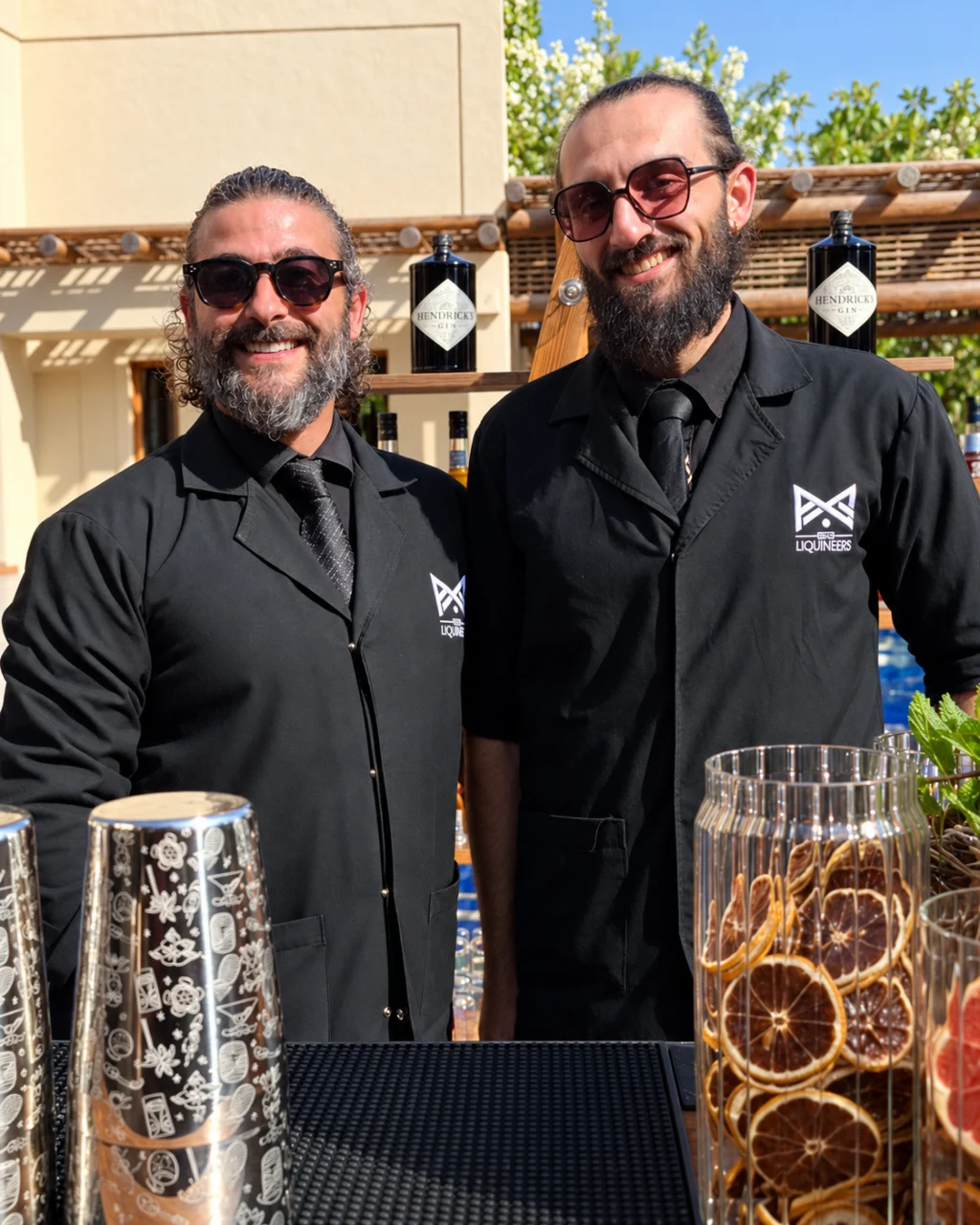 Tareq and Yamam smiling behind an outdoor bar in LiquiNeers uniforms, with cocktail tools and dried citrus garnishes