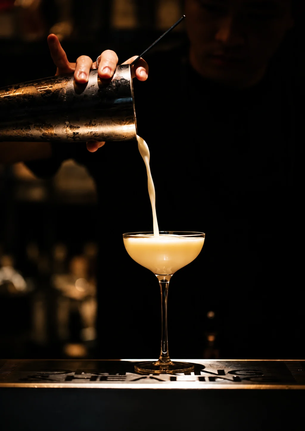 A creamy drink poured from a metal shaker into a coupe glass against a dark bar background.