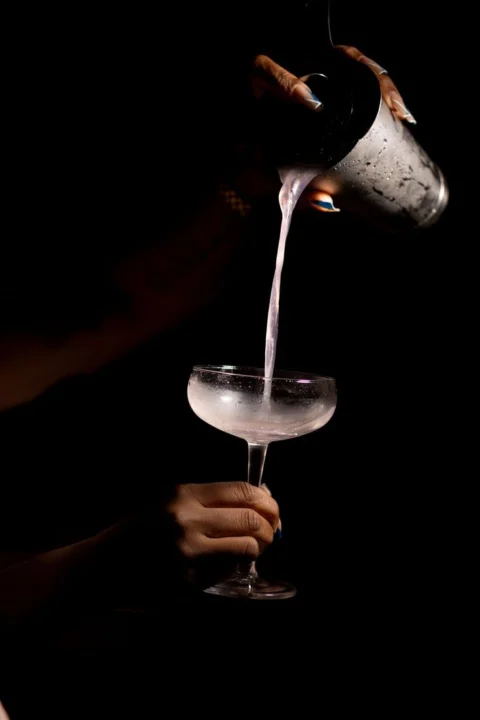 A pale pink drink poured from a cocktail shaker into a coupe glass against a dark background