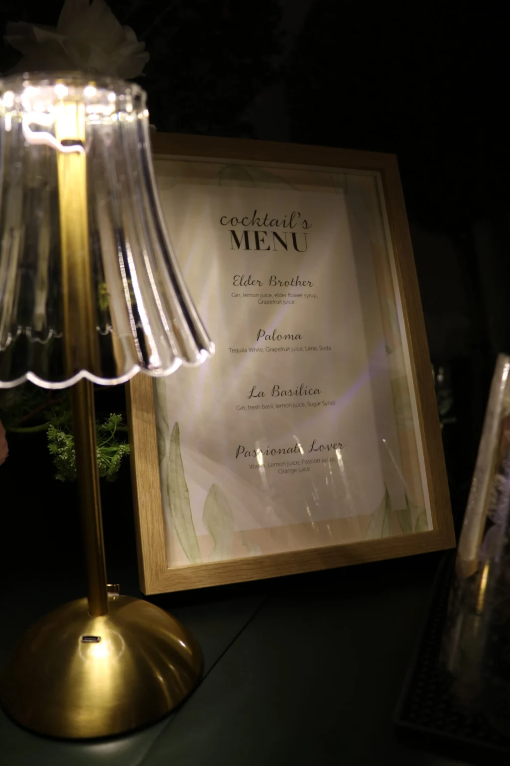 A framed cocktail menu beside an illuminated glass-shade lamp on a bar counter